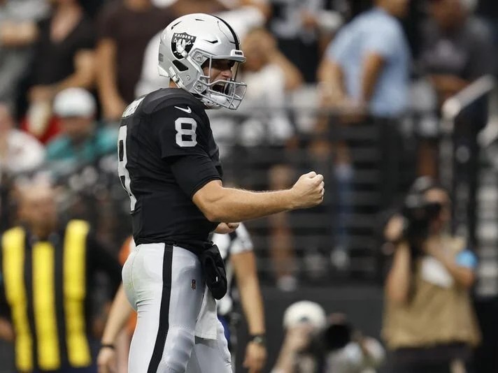Kirk Cousins stands on the field in a black Raiders jersey, number 8, clenching a fist after a play.