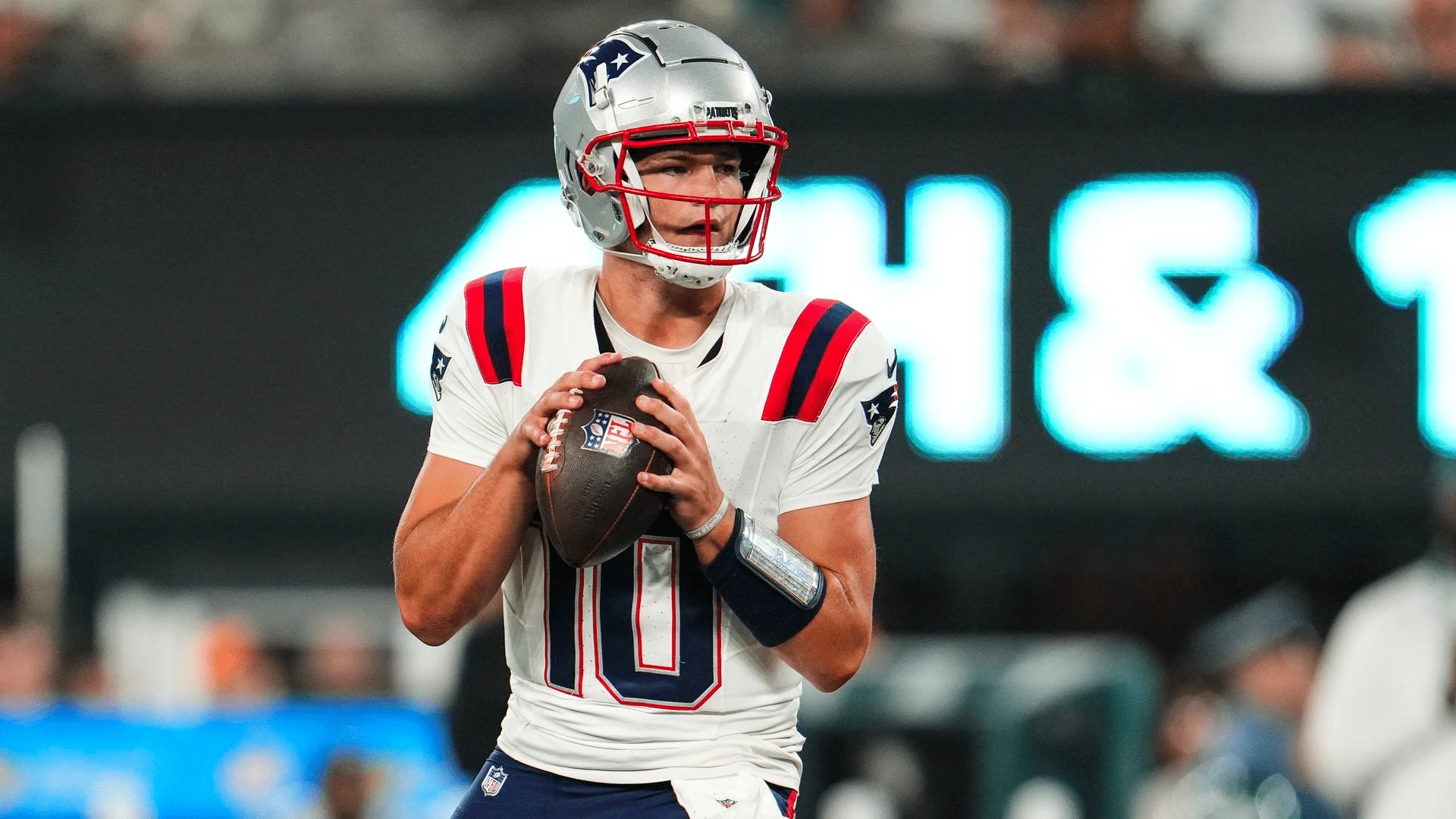 Drake Maye drops back to pass in a white Patriots jersey, holding the ball at chest height as he looks downfield.