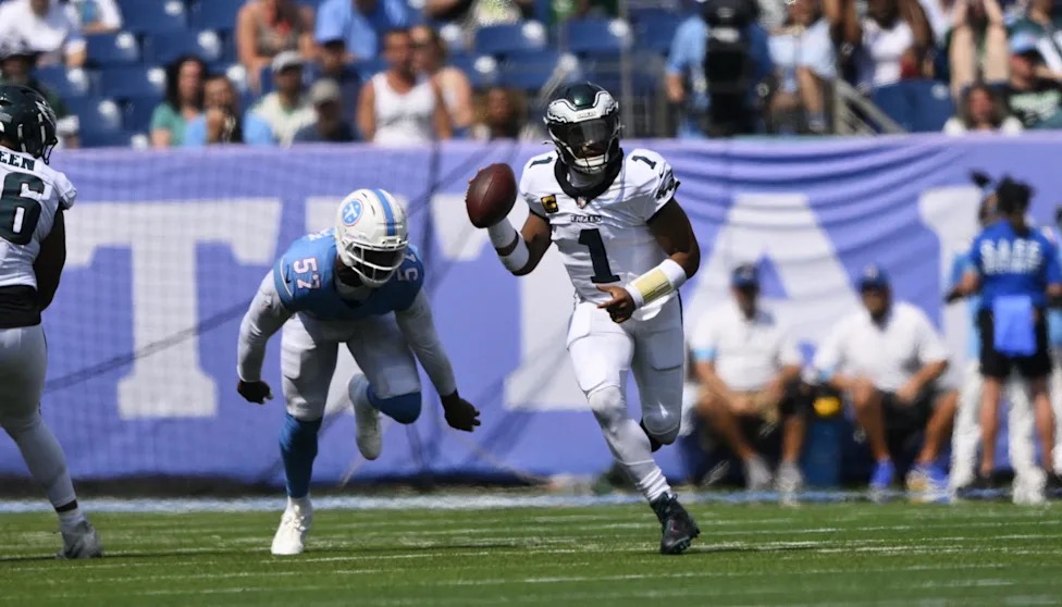 Jalen Hurts scrambles out of the pocket in a white Eagles jersey, a Titans defender chasing him down from behind.