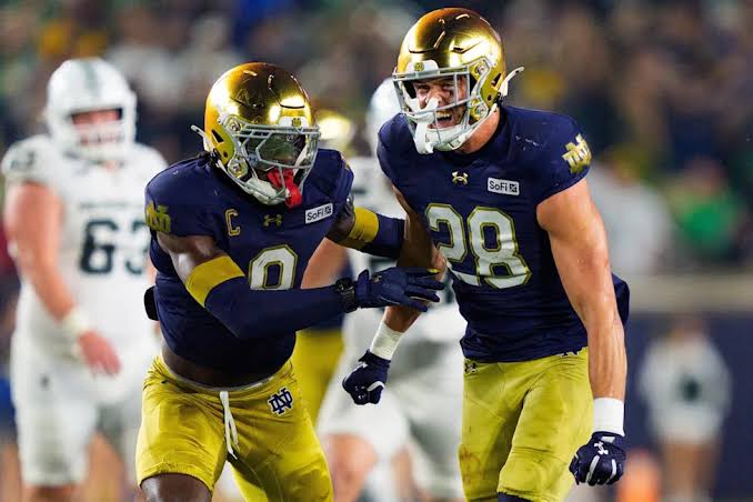 Two Notre Dame defenders in navy and gold celebrate a stop, one shouting as he runs off the field.