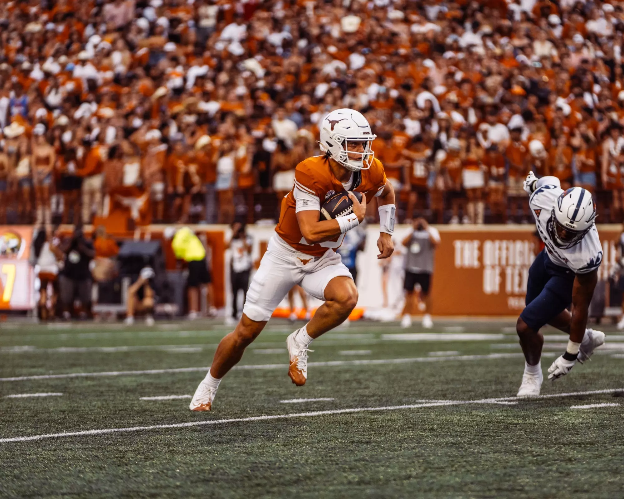 A Texas quarterback in burnt orange runs the ball upfield past a diving defender, a packed orange crowd behind him.